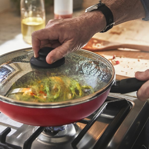 Person lifts lid of red frying pan on stove, revealing steaming food, demonstrating its non-stick and heat efficiency.