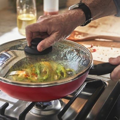 Person lifts lid of red frying pan on stove, revealing steaming food, demonstrating its non-stick and heat efficiency.