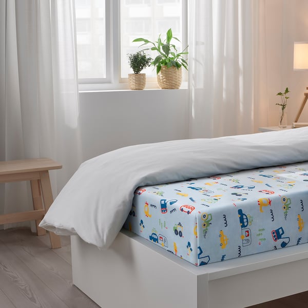 Childs bed with white vehicle-patterned sheet, white pillow, wooden bench, potted plants on windowsill.