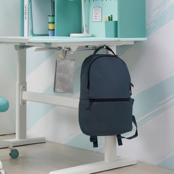 A grey BERGLÄRKA backpack hangs on a hook beside a desk with adjustable height and tiltable surface.