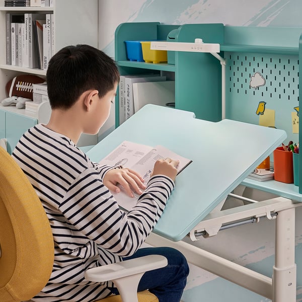 Boy sits at adjustable study desk, tilted table top, books nearby.