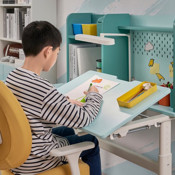 Child sitting at adjustable desk with tilted work surface, drawing. Desk has pegboard and shelves for storage.