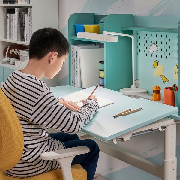 A boy writes at teal BERGLÄRKA desk with angled surface, lamp, and colourful containers.