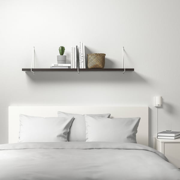 Minimalist bedroom with white BERGSHULT wall shelf holding books, plant, and basket. Bed with white bedding below.