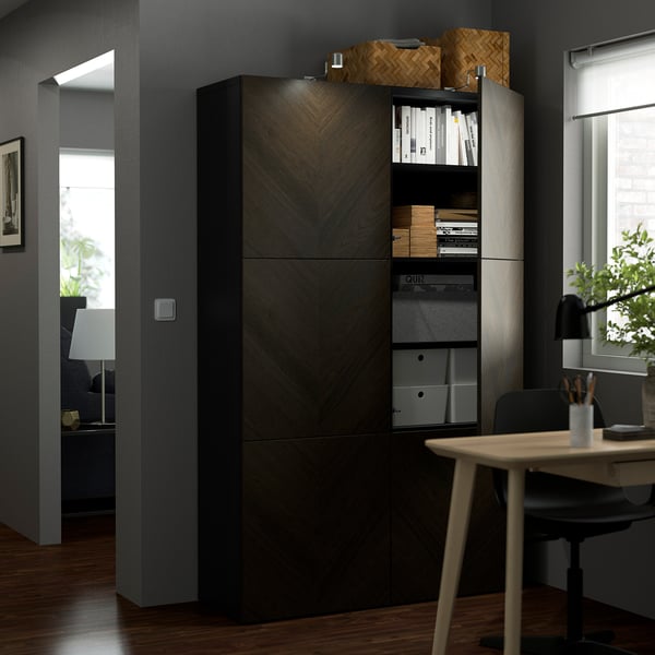 Modern office with dark, tall BESTÅ storage unit, open doors revealing shelves with files and bins, desk with chair, lamp, and plants.