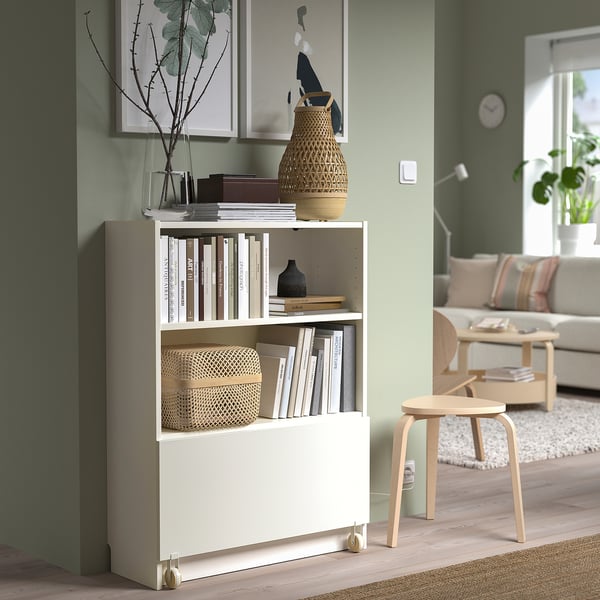 A white BILLY bookcase with a drawer, filled with books and decorated with vases, next to a light wooden stool and sofa in a cosy living room.