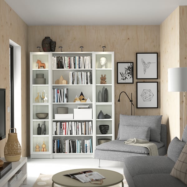 Modern living room with white BILLY bookcases, grey sofa, and decorative art frames. Wooden walls, glass doors, and various ornaments.