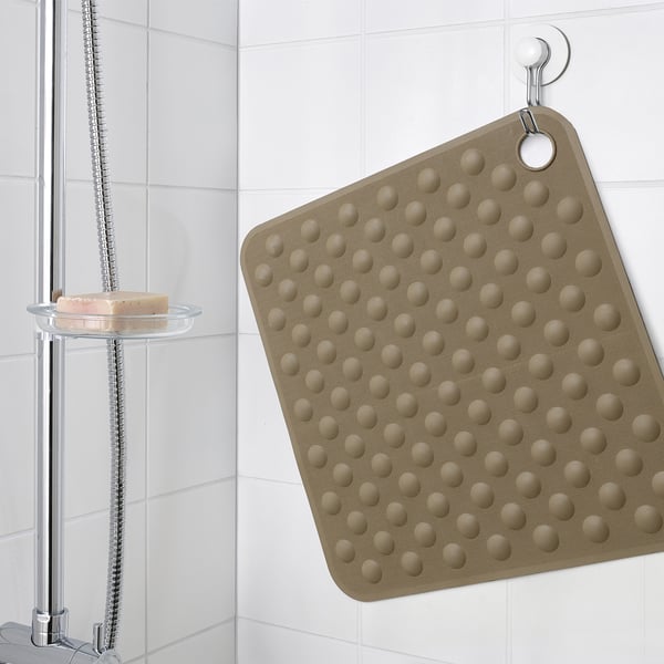 A beige bath mat hangs on a hook in a shower, featuring suction cups on its textured surface.
