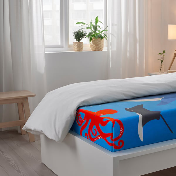 Bright bedroom with a blue bed sheet showing red octopus and grey fish, white pillows, plants by the window, wood floor, light curtains.