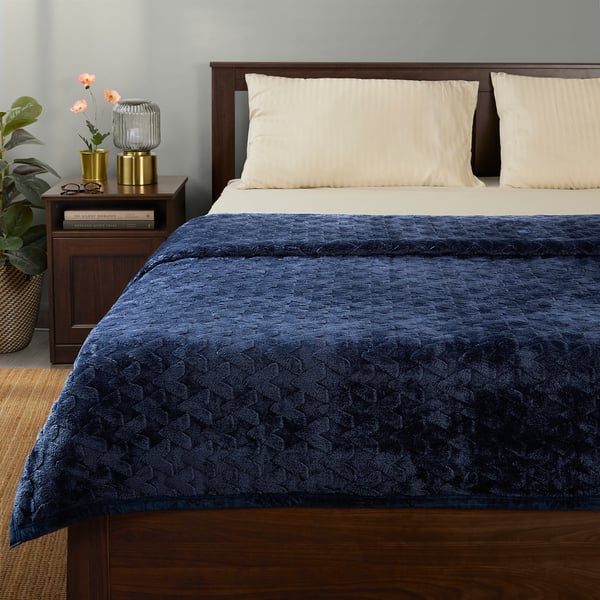 Navy blue BLIXABACKEN blanket on bed with beige pillows, geometric pattern. Recycled polyester, with small plants and lamp nearby.