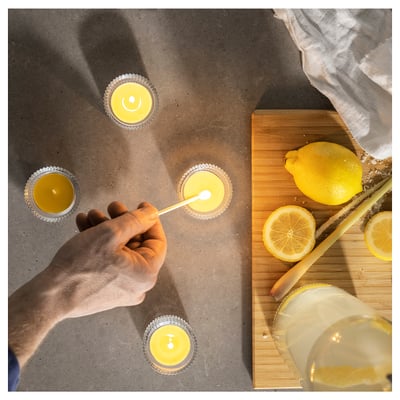 Person lights lemon-scented candles. Four yellow candles in metal holders; one lit. Lemons, knife, cloth on wooden board.