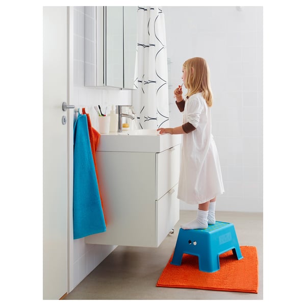A child stands on a blue step stool in a bathroom, brushing their teeth with a white sink visible.