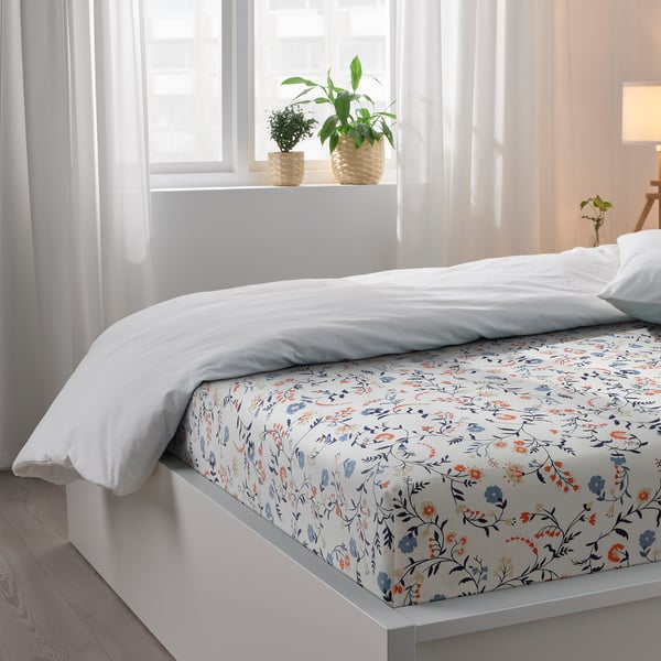 Bright, floral-patterned flat sheet and white pillows on a cosy bed, with green plants on the window sill, creating a serene, inviting bedroom.
