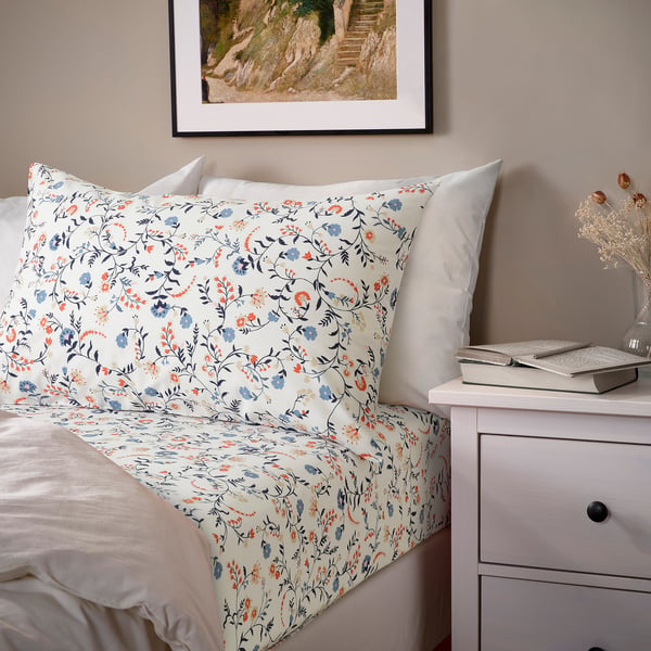 Bed with floral BRUDKRUSBÄR white cotton sheet, matching pillowcases, plus side table with laptop and flowers.