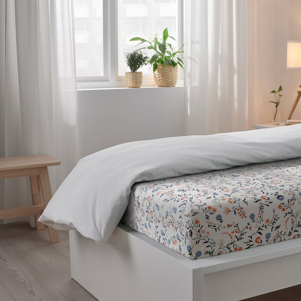 Bright bedroom with white bed, floral sheet, two plants on the sill, natural light.