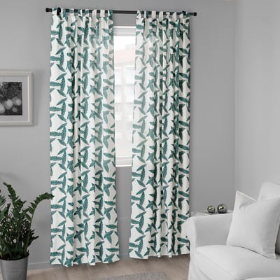 White curtains with leaf patterns hang over a window, appearing tall and wide, covering it almost entirely.