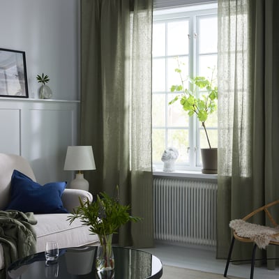 Modern living room with large olive green linen curtains behind a cosy chair with blue and white pillows, a white lamp, and a vase with greenery.