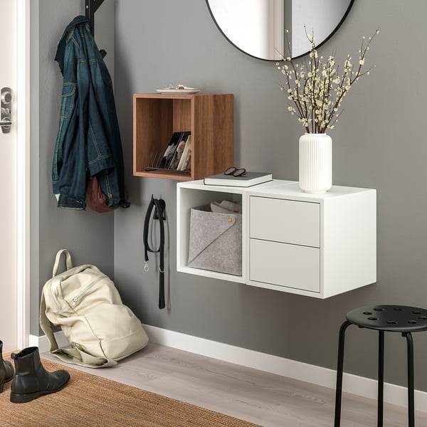 Modern entryway with wall-mounted white EKET storage units. Shelves with various items, a round mirror, a stool, and coat hooks.