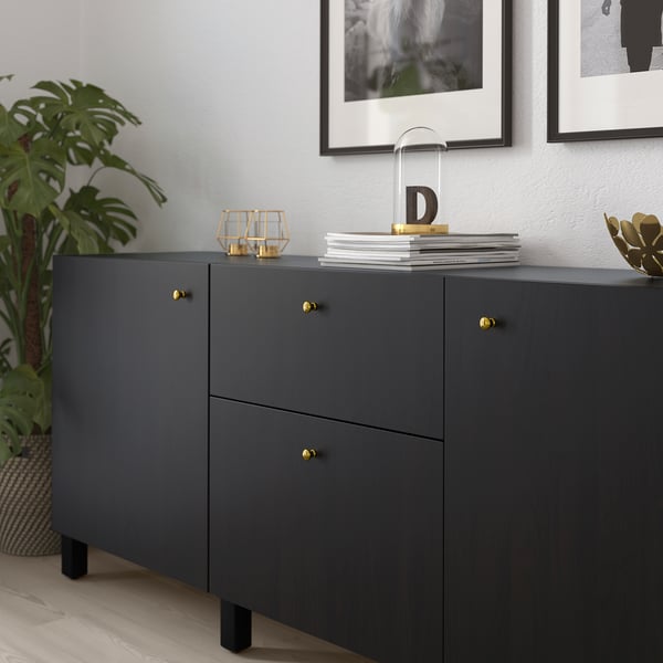 Dark sideboard with gold handles and knobs, topped with books and decorative items, placed near a large green plant.