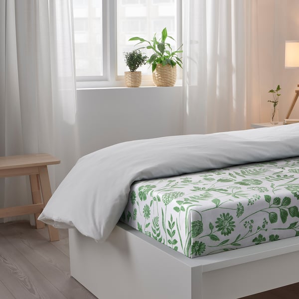 Bedroom scene with white bedding, green floral sheet, plants on windowsill.
