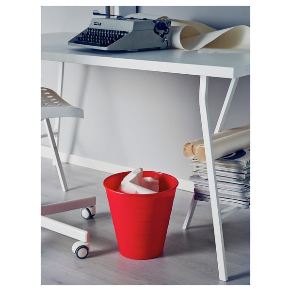 A red plastic bin on wheels sits below a white desk with a typewriter and paper rolls.