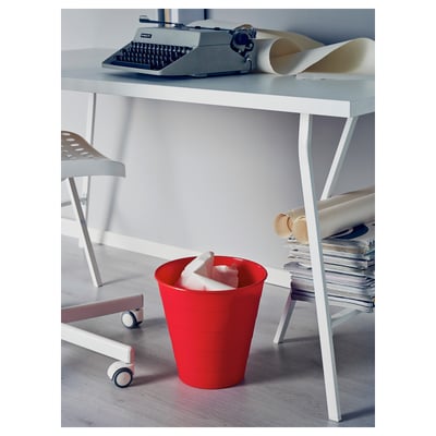 A red plastic bin on wheels sits below a white desk with a typewriter and paper rolls.