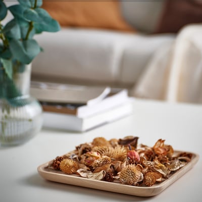 Small wooden tray named FRÖFINK, holding dried flowers, placed near a white sofa with a stack of books and a green plant.