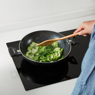 A person stir-fries vegetables in a two-handled non-stick kadai wok using a wooden spatula.