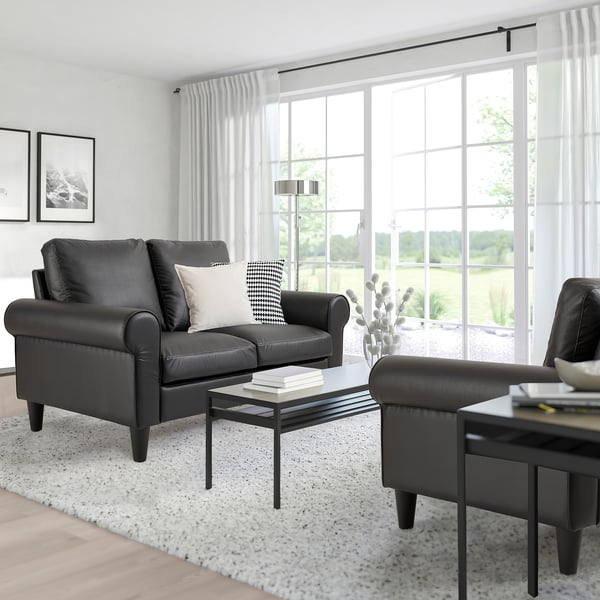 Modern living room with grey sofa, black coffee table, and ottoman. Large windows with sheer curtains. Minimalistic style, white & black accents.