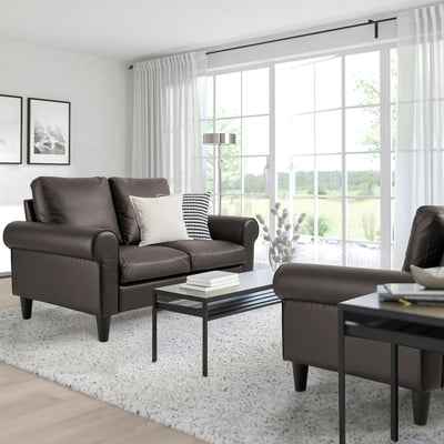 Modern living room with GAMMALBYN leather sofa, ottoman, glass coffee table, and large window with curtains. Light rug enhances space.