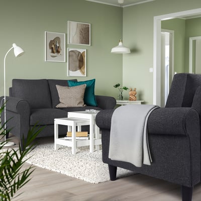 Modern living room with grey sofa & armchair, black/white pillows, blue cushion, green walls, white table & rug, hanging lamps, art, plant.