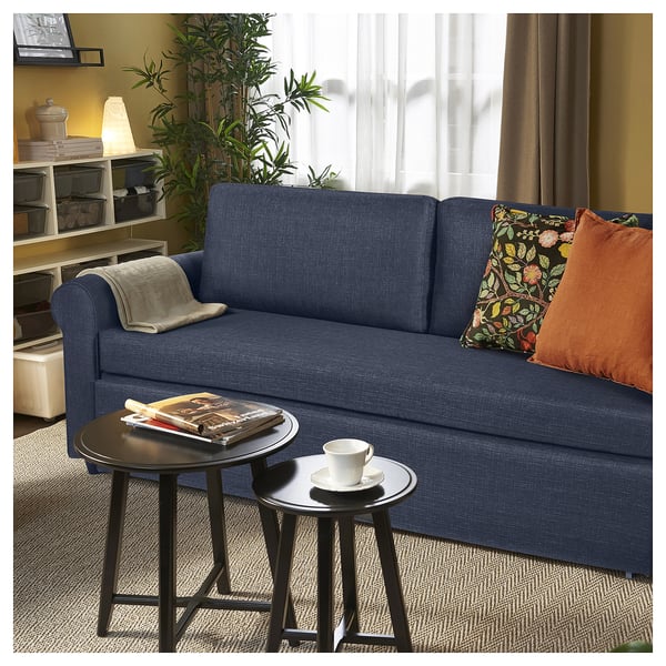 Modern living room with blue GAMMALBYN sofa-bed, cushioned seating, accent pillows, small round side tables, books, cup, and green plant.