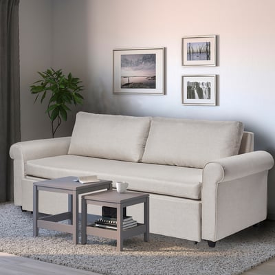 Small, light beige GAMMALBYN sofa-bed against neutral wall, flanked by framed art and plant. Accented by compact side table with books and cup.