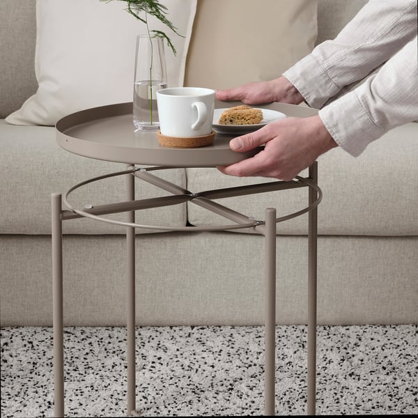 A person holding a tray with a coffee cup and biscuit from a GLADOM side table.