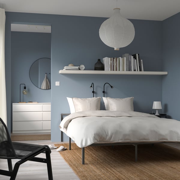 Minimalist bedroom with white bed, blue wall, light, books, mirror, and chair.