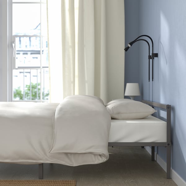 Bed frame under window, white bedding, lamp, curtains.