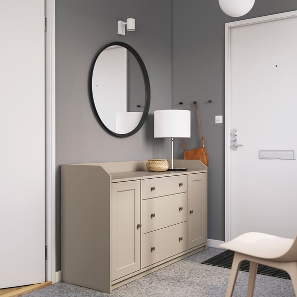 Contemporary entryway grey HAUGA sideboard, round black-framed mirror, white lamp, and beige chair, set against grey walls.
