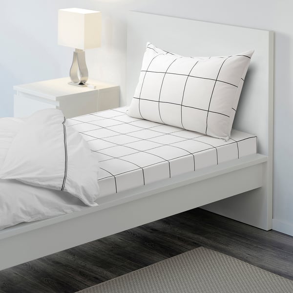 Bed with JÄMTSTARR sheets, white wall, bedside table, lamp, dark rug.