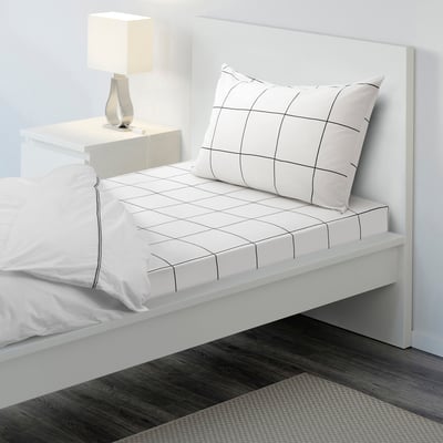 Bed with JÄMTSTARR sheets, white wall, bedside table, lamp, dark rug.