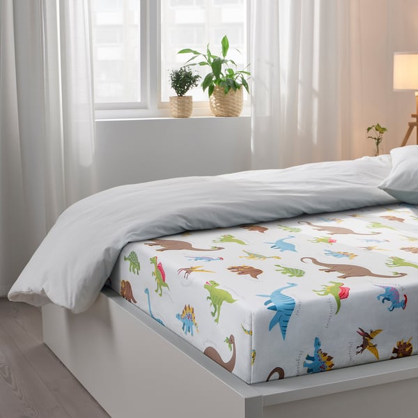 Childs bed with dinosaur-themed sheet; white curtains, plants.