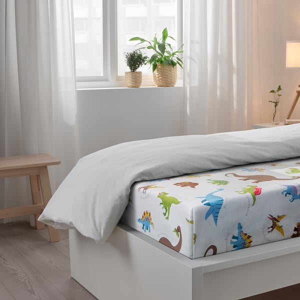 Bed with dinosaur-themed sheets, white pillows, wooden bench, and potted plants on windowsill.