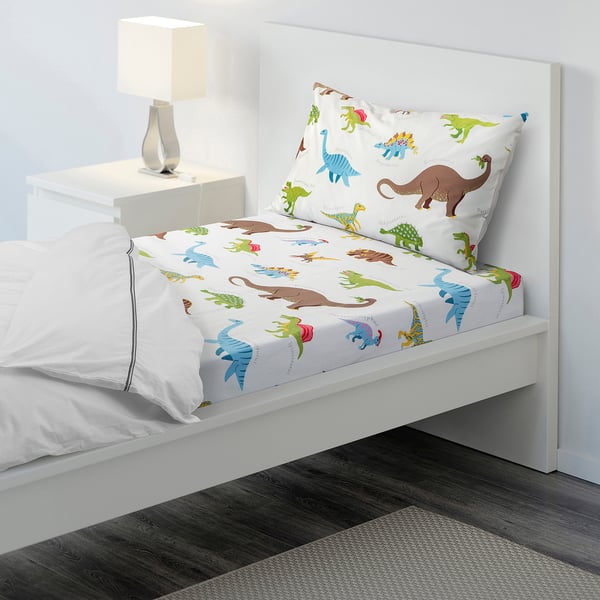 Dinosaur-themed bedding set in white with soft cotton blend, including fitted sheet and pillowcase, next to a bedside table with lamp.