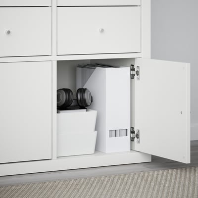 White KALLAX storage unit with open door showing stacked boxes and dumbbells.