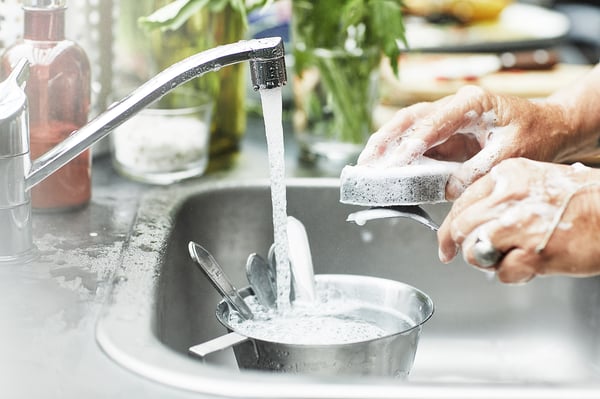 Person washing utensils in stainless sink. Water running from white tap, soap suds visible. Hands holding utensils and sponge.
