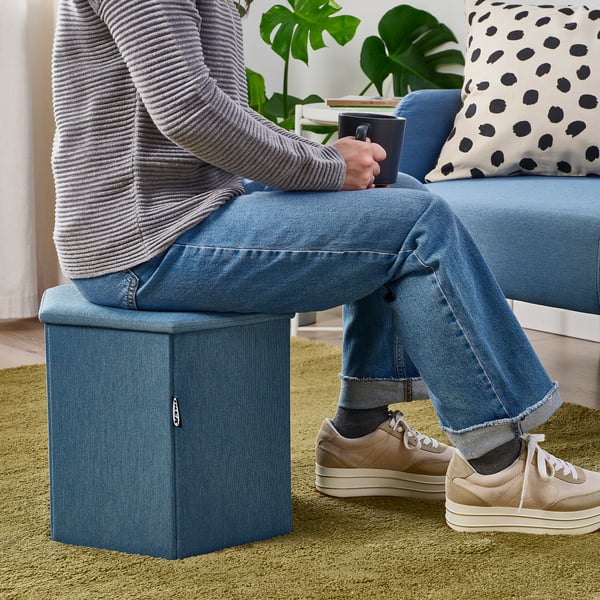 Person sits on blue KJUGE, footrest raised, holding mug. Leopard pillow on blue couch. Green carpeted floor.