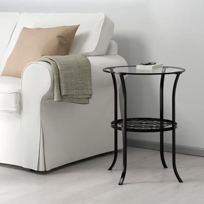 KLINGSBO black metal side table sits beside a white sofa, featuring curved legs and a glass top. Its small, elegant design holds items neatly.