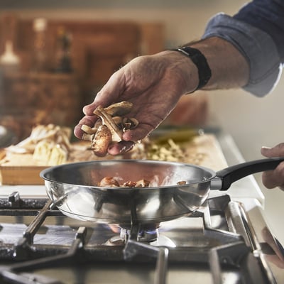 A person holds fresh spices over a sizzling stainless steel frying pan on a stove, demonstrating cooking preparation.