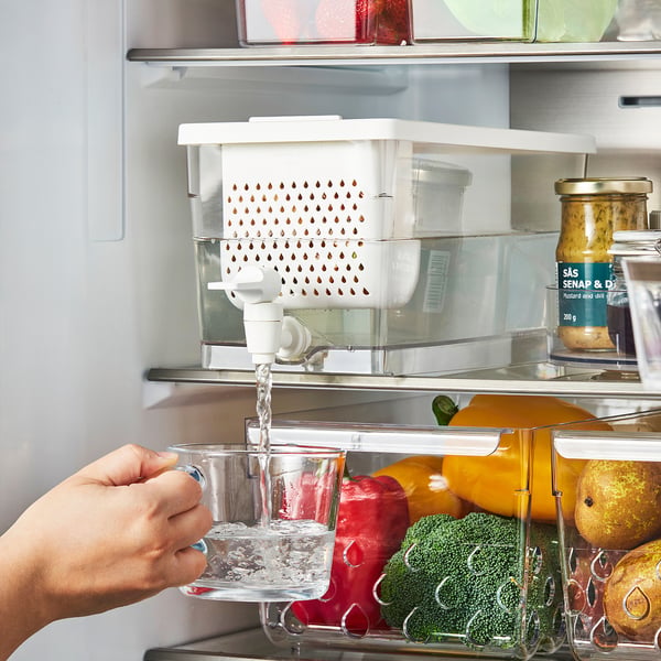 Person pouring water from a fridge dispenser into a glass. The dispenser is connected to a container filled with vegetables in the fridge door.