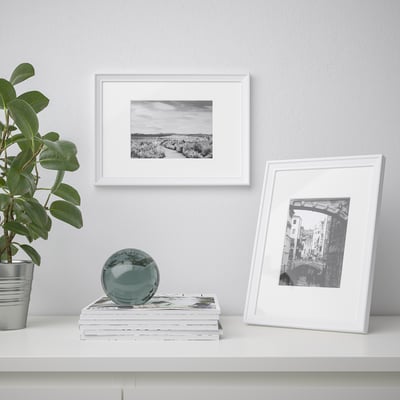 A white shelf with a plant, books, and two picture frames. One frame is upright displaying a bridge photo, the other hangs showing a landscape.