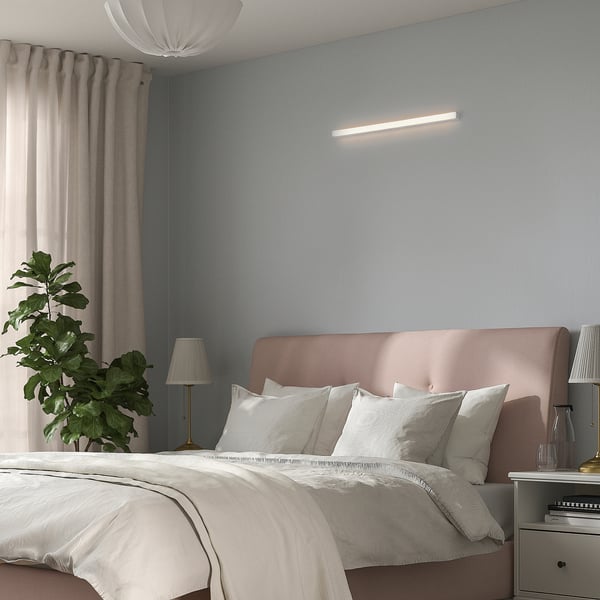 Minimalist bedroom with lit led light above bed, white bedding, pillows, plant, lamp, curtains, and bedside table with accessories.